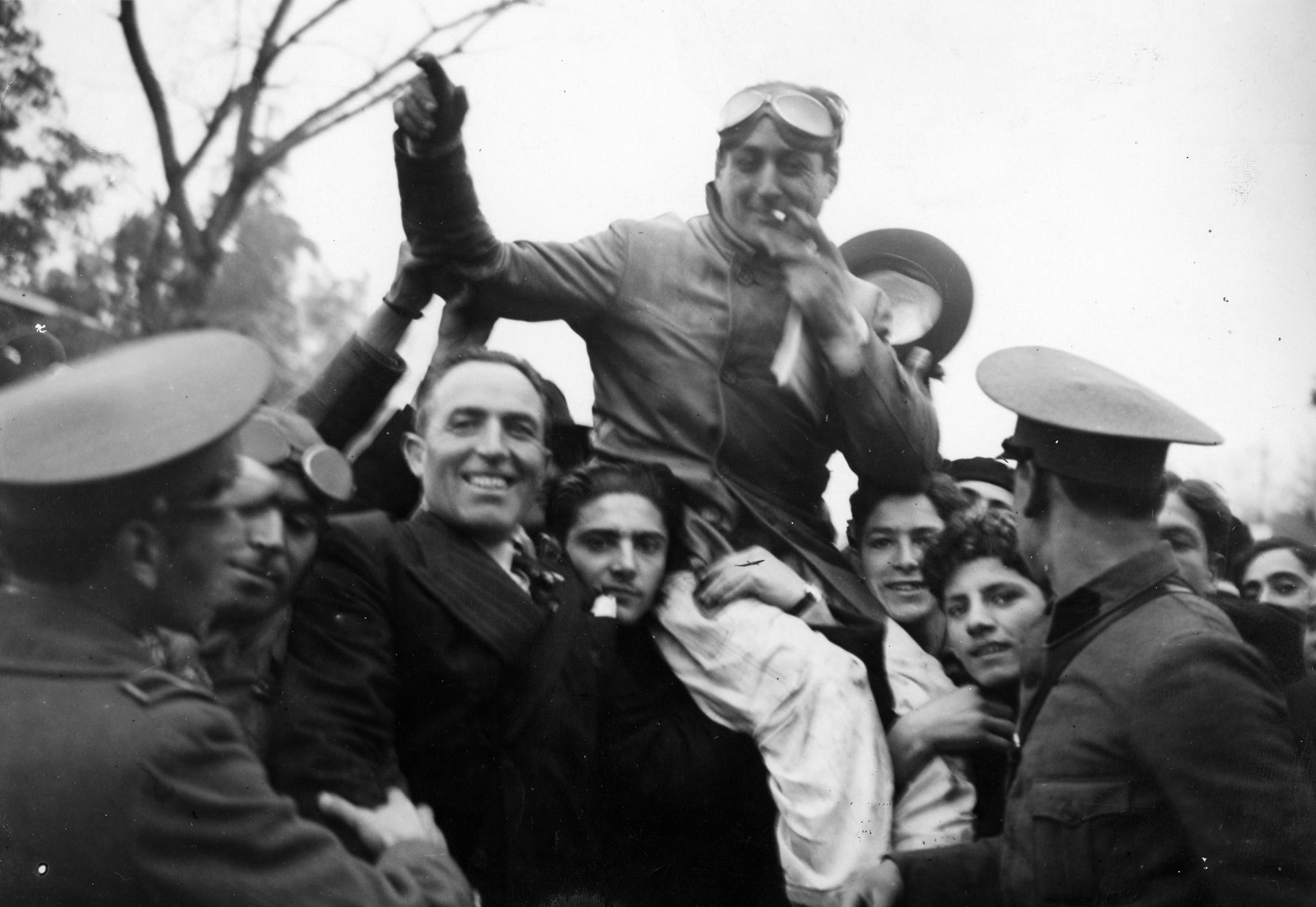A driver in a leather jacket, goggles pushed up on his forehead and a cigarette in his fingers at his mouth, is carried shoulder-high through a cheering crowd of young men, one gloved fist raised; two soldiers in peaked caps stand in the foreground.