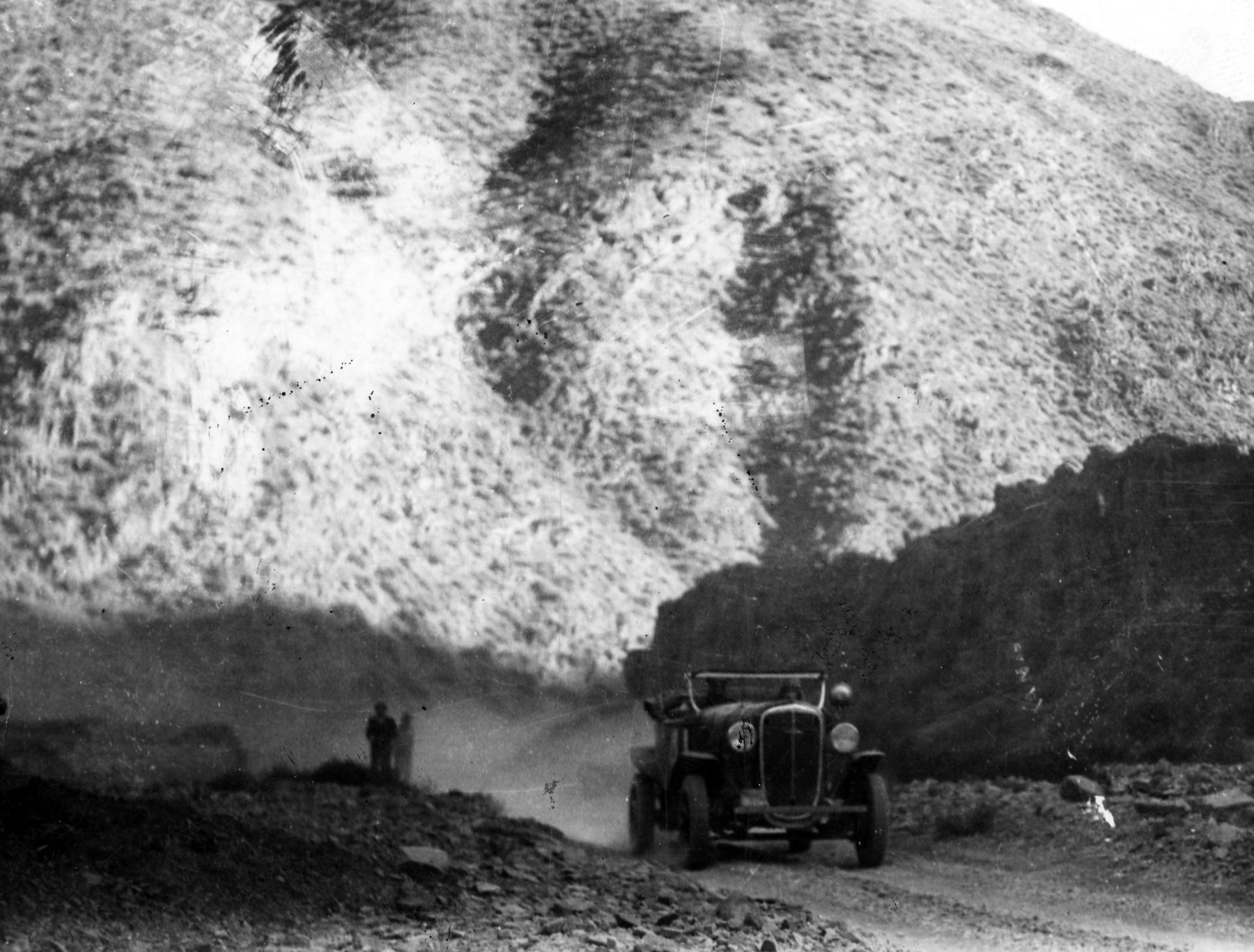 A lone open touring car climbs a gravel road at the foot of a huge, pale mountainside streaked with dark scrub, two figures standing by the road further back.