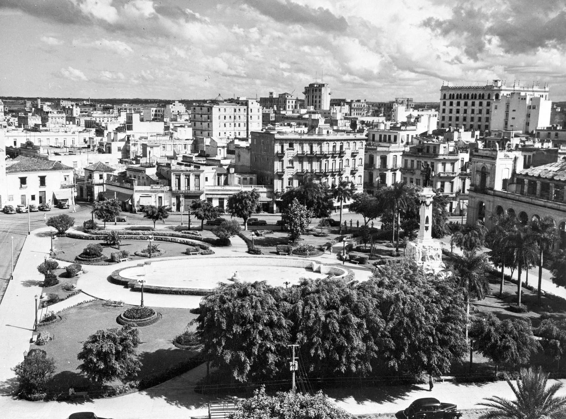 A high view over a city square: a laid-out park with a round basin, clipped hedges, lamp standards and palms, a statue of a standing man with an outstretched arm on a tall column at the right, parked late-1940s cars along the street, and apartment blocks and hotels behind.