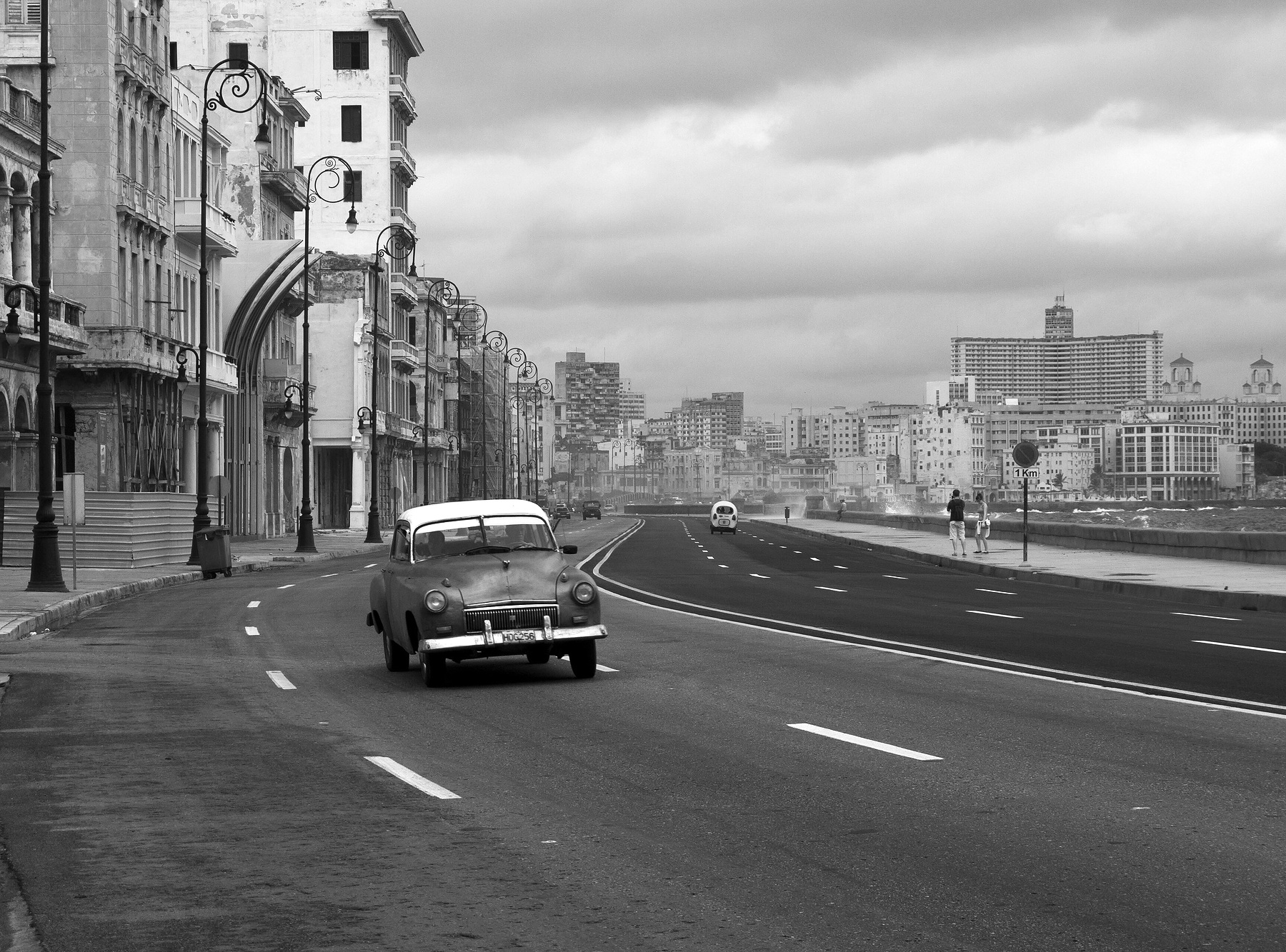 A 1950s saloon car drives along a wide seafront road on a stormy day, spray over the sea wall at the right, colonnaded, weathered buildings and iron lamp-posts at the left and towers on the skyline.