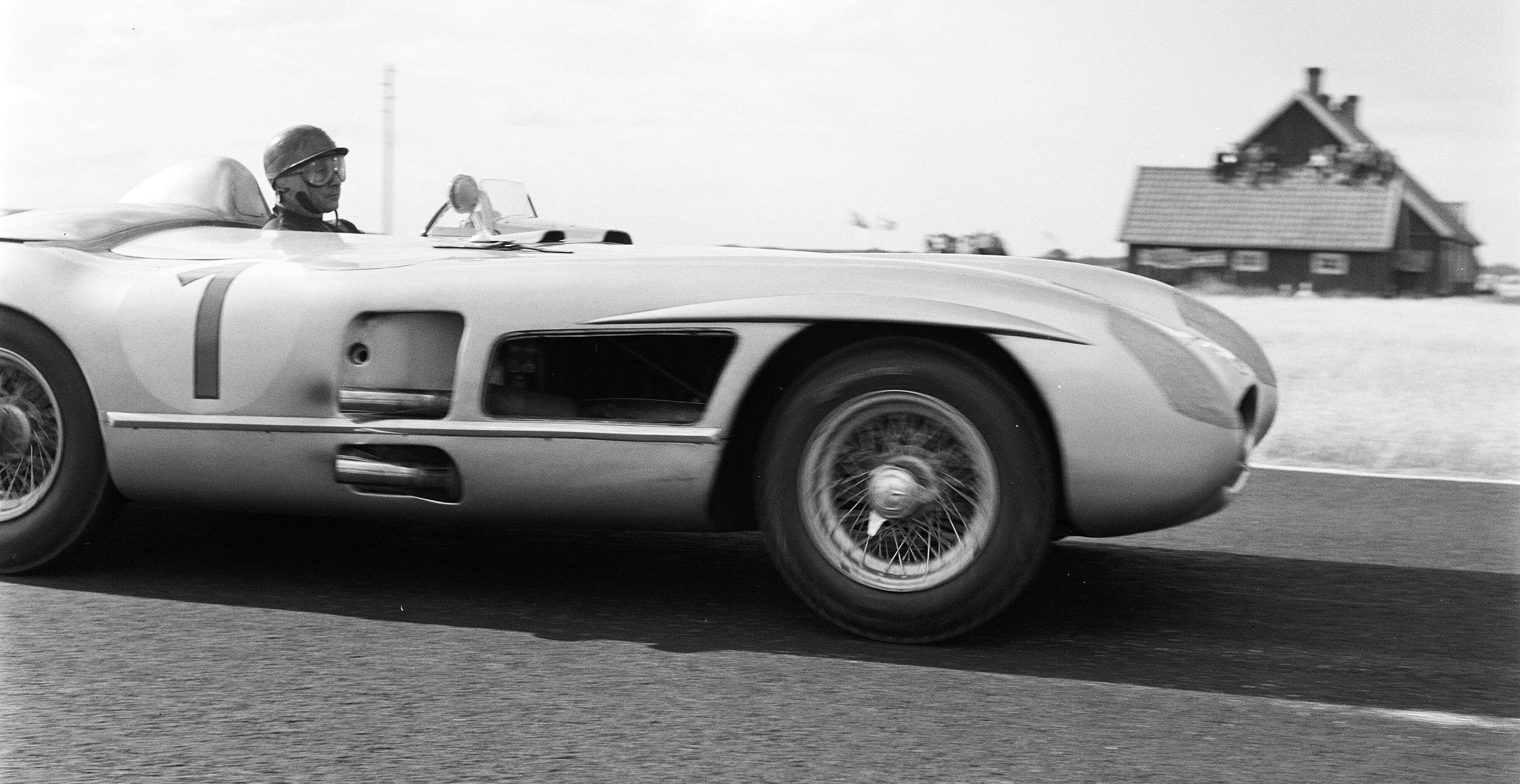 A panned side view: an open sports-racing car numbered 1 passes close to the camera, its driver in a dark peaked helmet and goggles; a farmhouse with a steep roof blurs past behind under a pale sky.