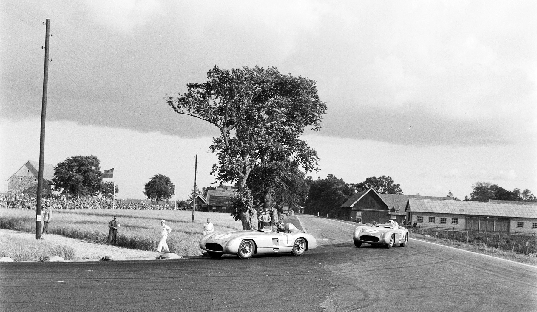 Two low open sports-racing cars round a bend beside a tall lone tree and farm buildings; marshals in white stand at the edge of a field, spectators and a flag beyond.