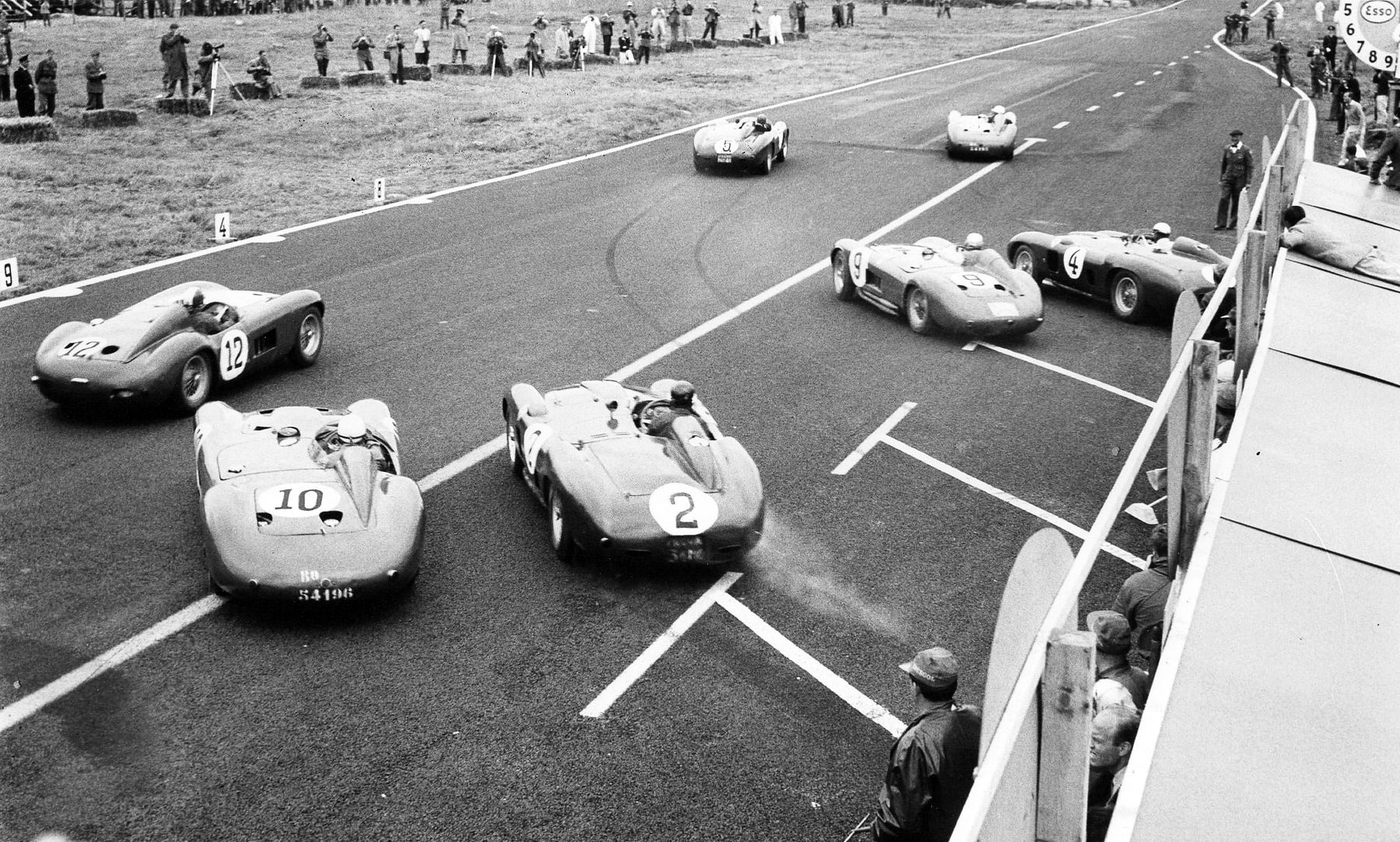 A high view of a start on a wide straight marked with white lines: sports-racing cars numbered 12, 10 and 2 pull away in the foreground, 9 and 4 further on at the right beside the pit rail and two more ahead; photographers and spectators line the grass verge, and men watch from behind the rail.
