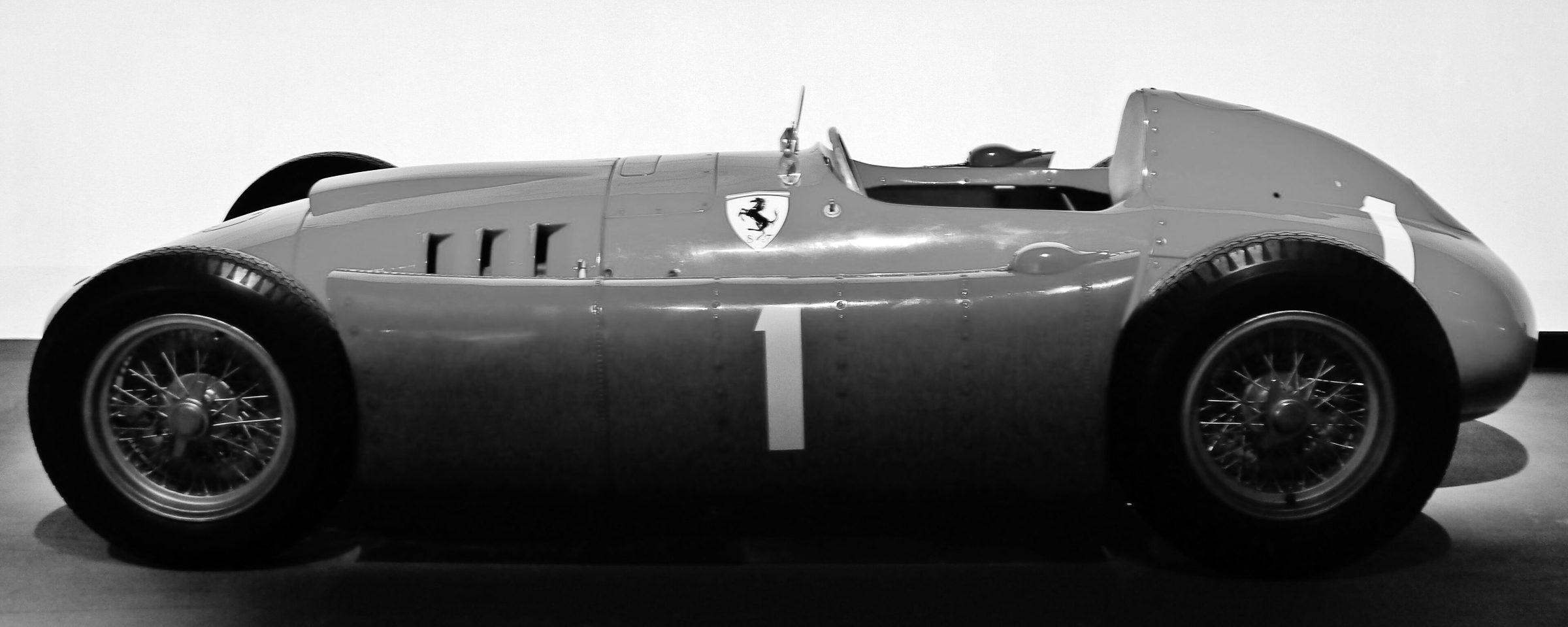 A single-seater seen exactly side-on, its nose to the left, on a dark floor against a pale wall: a smooth riveted body swelling between the wire wheels, a prancing-horse shield and three louvres on the flank, a large 1 on its side and a headrest behind the open cockpit.