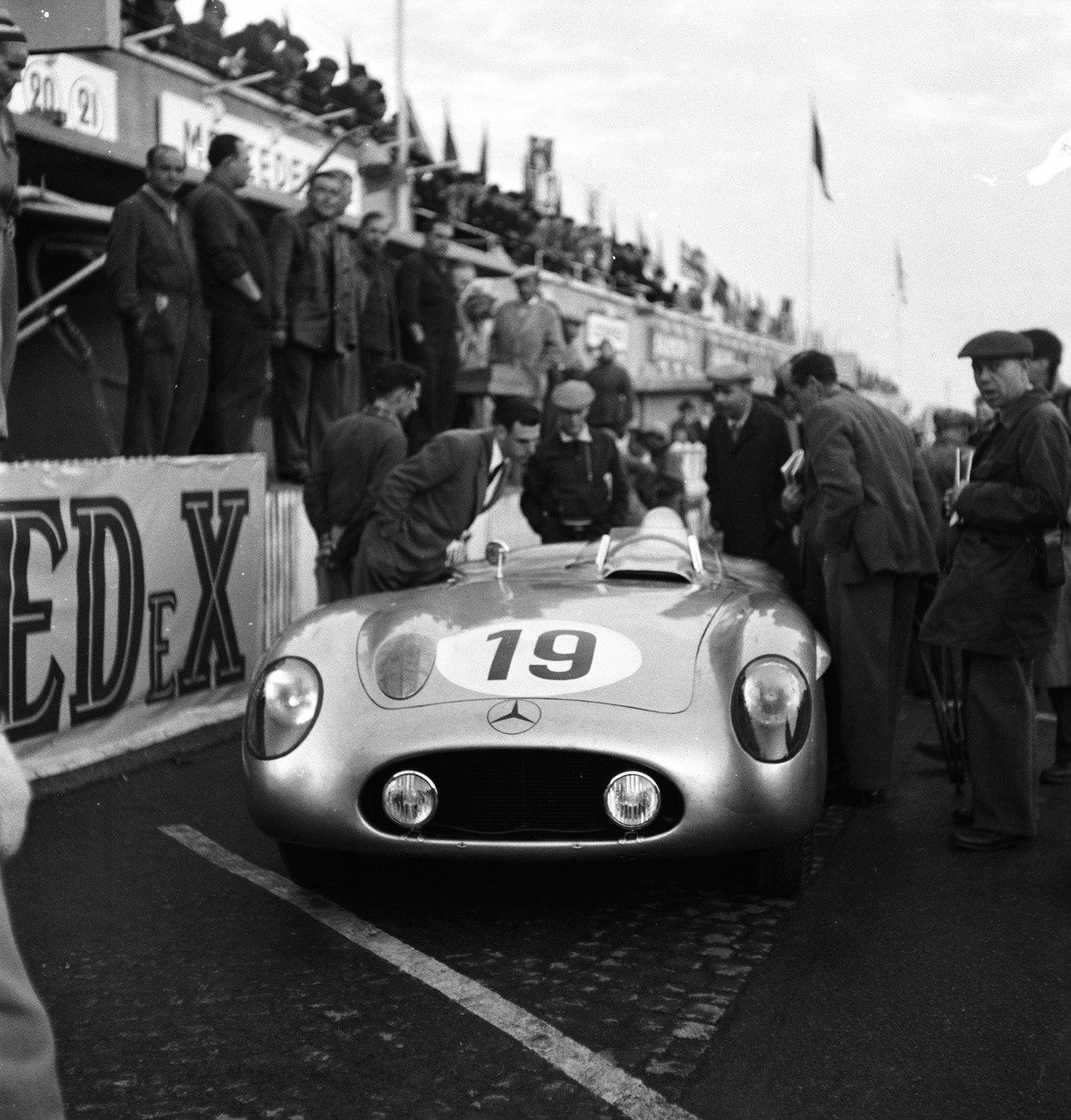 Head-on in the pits, a low open sports-racing car numbered 19, the three-pointed star on its nose, surrounded by mechanics and men in caps and coats; a hoarding at the left and the pit balconies behind.
