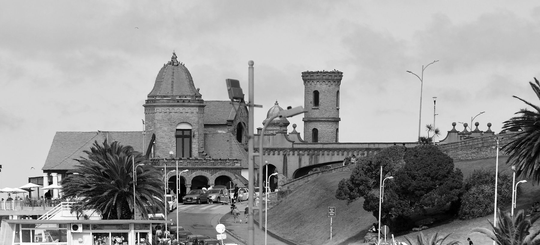 A mock-medieval pavilion with a tiled dome and a crenellated tower on a grassy bluff above a coast road, with palms, street lamps, parked cars and a few people below.