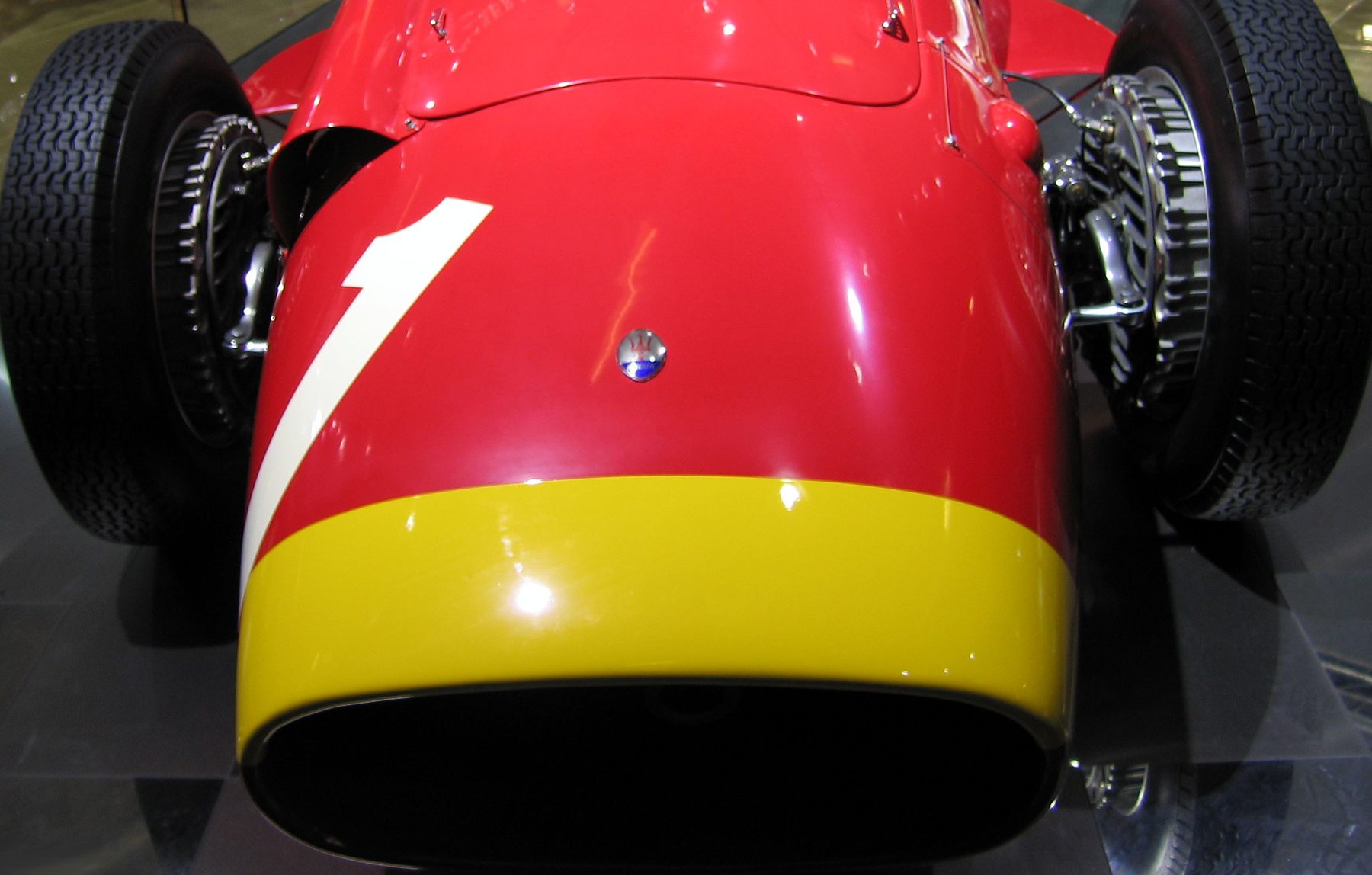 In colour: a red Maserati 250F seen head-on, a white 1 swept across its nose above the trident badge, a broad yellow band round the mouth of the air intake and a front wheel to either side, on a polished showroom floor.