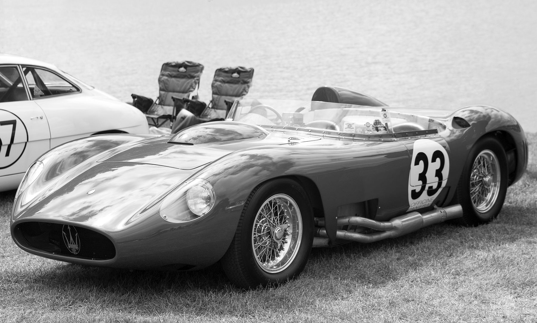 A sports-racing car with long, rounded bodywork, a roundel with the number 33 on its flank, wire wheels and side exhausts, parked on a lawn by the water; the flank of another car and two folding chairs at the left.
