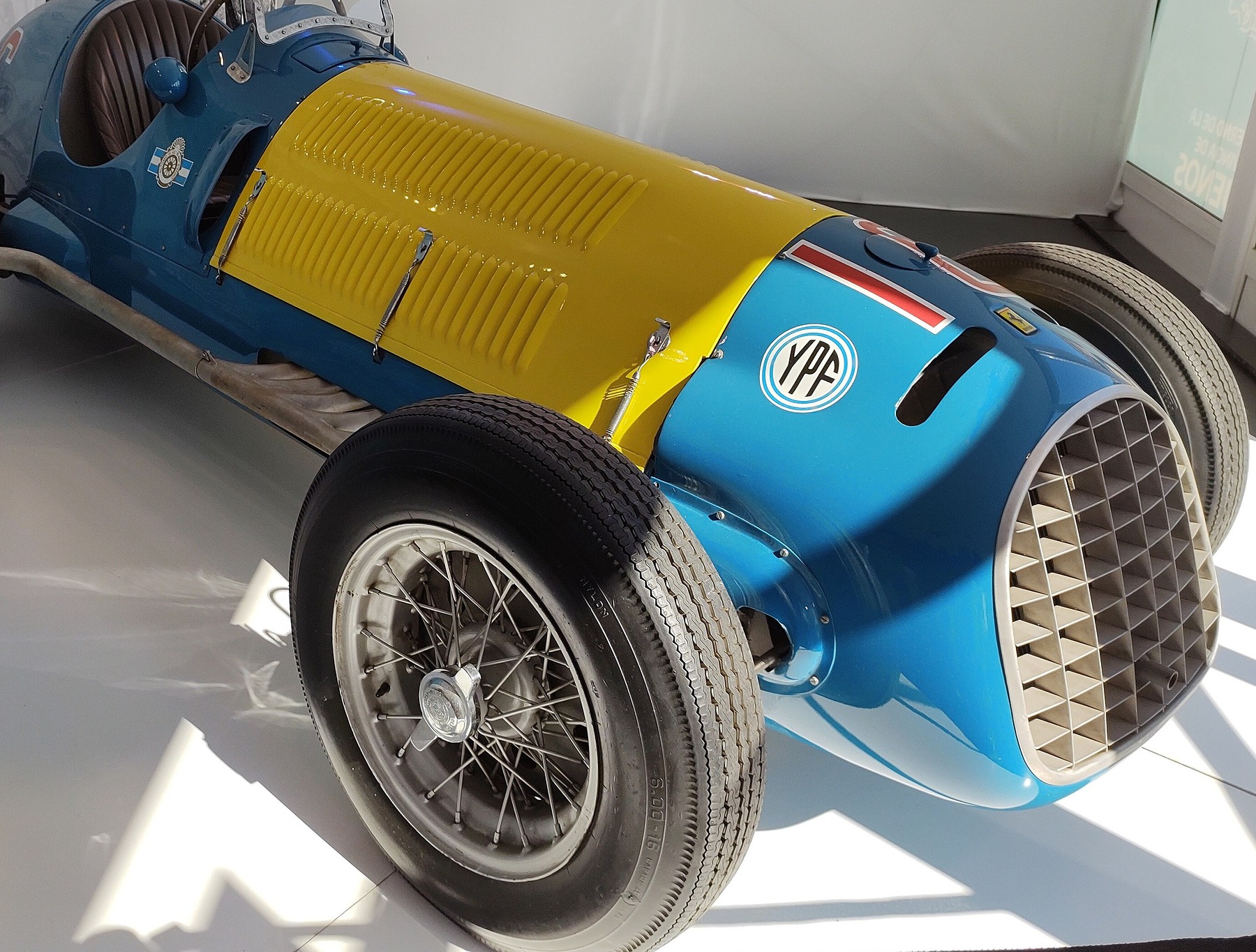 In colour: an open-wheeled racing car on a white floor, its body sky blue and its long louvred bonnet yellow, a YPF roundel, a red stripe and a small badge on the nose, an egg-crate grille and wire wheels.