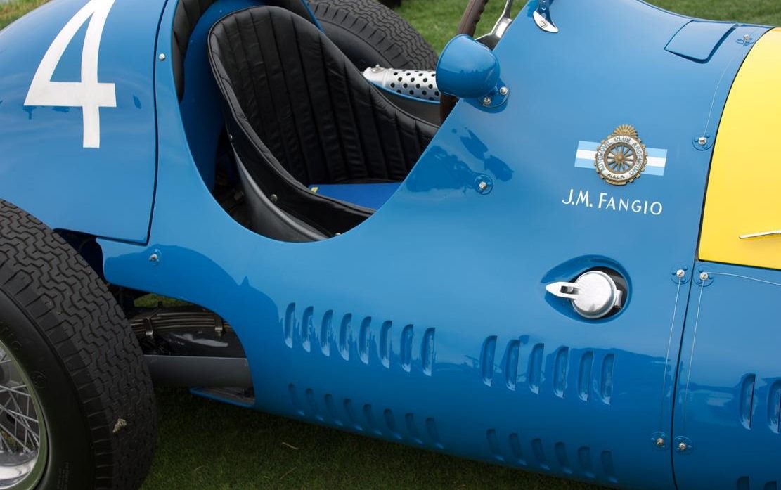 In colour: close on the sky-blue flank of a racing car, a white number 4 on the tail, a club badge between two small pale blue and white flags, the lettering J.M. Fangio beneath it, louvres, a fuel filler cap and the edge of a yellow bonnet.