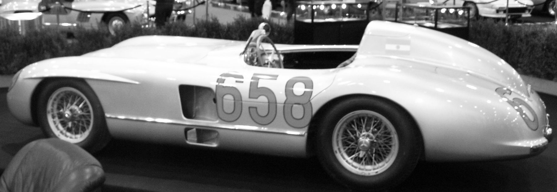 A two-seater sports racing car in profile, nose to the left, with 658 painted large on its door and again on its tail, a small flag of Argentina on the head fairing, a wooden-rimmed steering wheel and wire wheels, on a black plinth; the back of an armchair in the foreground and legs and display cases behind.