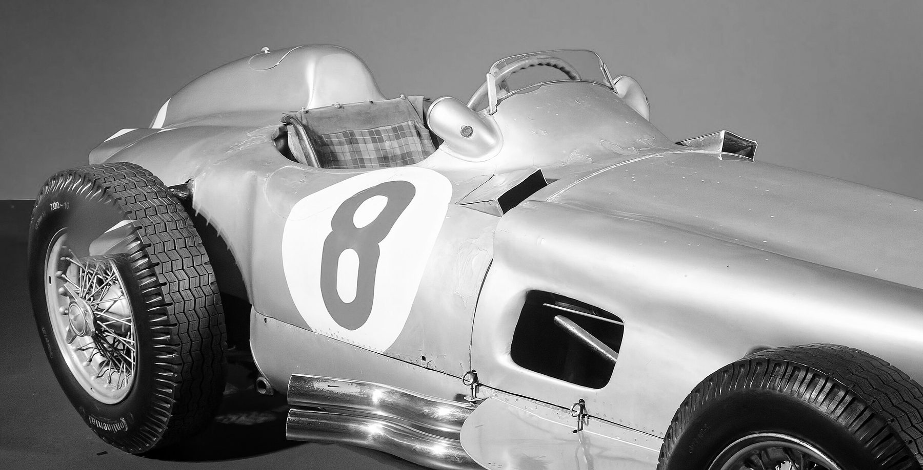 The cockpit of a Mercedes-Benz W196 open-wheeler, close: a wrap-round windscreen, a tartan-covered seat and a headrest fairing, and a large 8 in a roundel on the flank, against a plain wall.