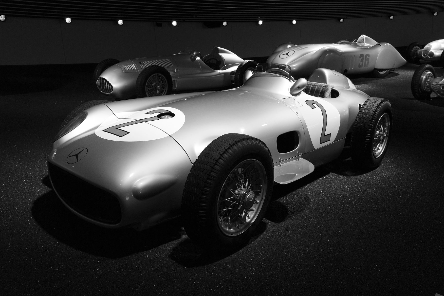An open-wheeled Mercedes-Benz W196 with the number 2 in roundels on its nose and flank and the three-pointed star on its low nose, on a dark floor; pre-war racing cars stand behind it.