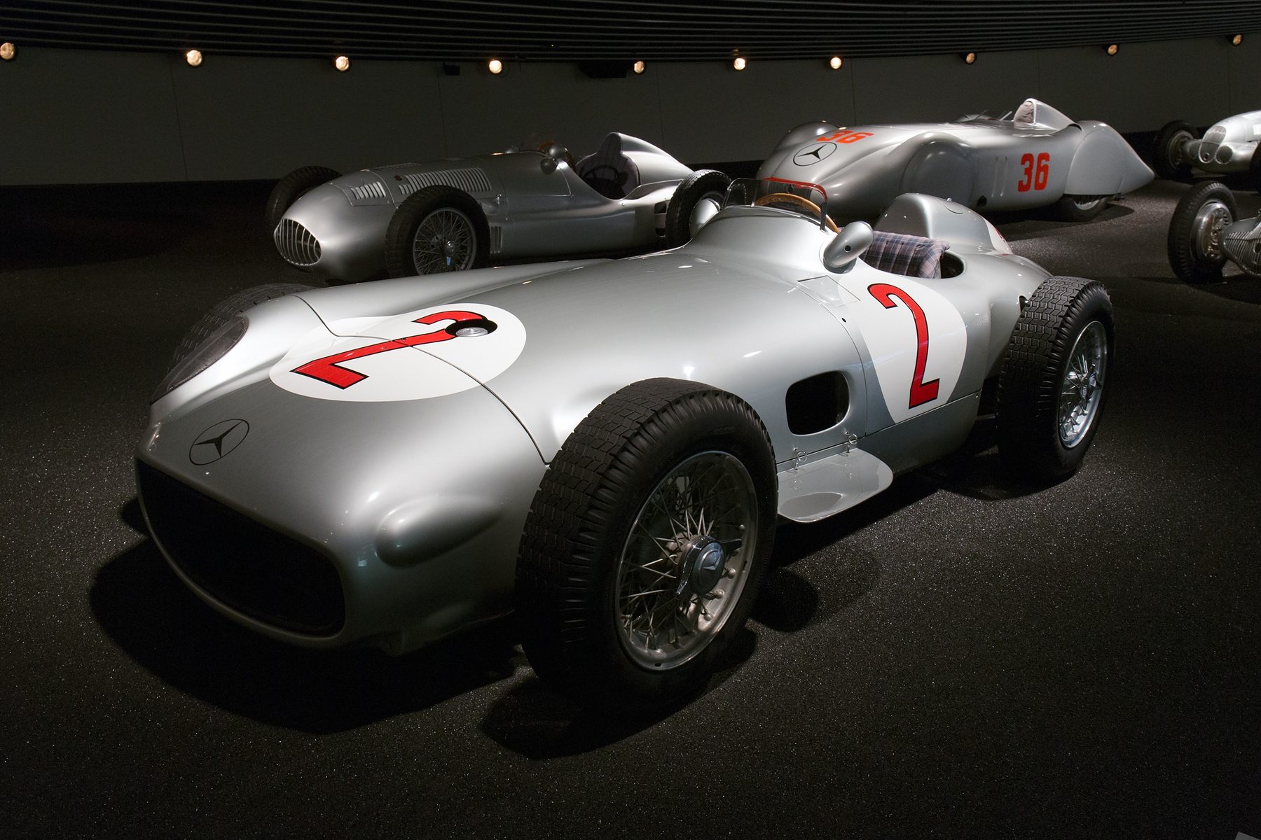 In colour: a silver Mercedes-Benz W196 open-wheeler with a red number 2 in white roundels on its nose and flank, the three-pointed star on the low nose, wire wheels and a tartan seat, on a dark floor; three pre-war silver racing cars stand behind it, one numbered 36.