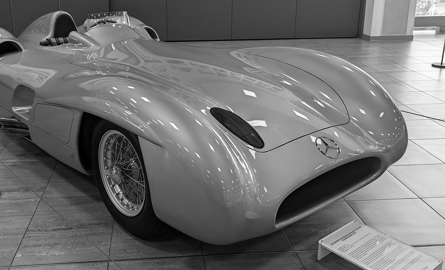 The streamlined body of a Mercedes-Benz W196 seen from the front three-quarter on a tiled showroom floor: a wide oval mouth, the three-pointed star, the front wheel enclosed in a flowing wing, a small curved windscreen and a headrest fairing; an information card stands at its nose and a rope line behind.