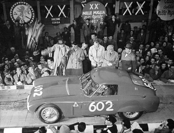A low closed sports-racing car numbered 602 on a start ramp; officials in raincoats and hats look down at it, spectators are packed behind, and banners with the race’s name hang above.