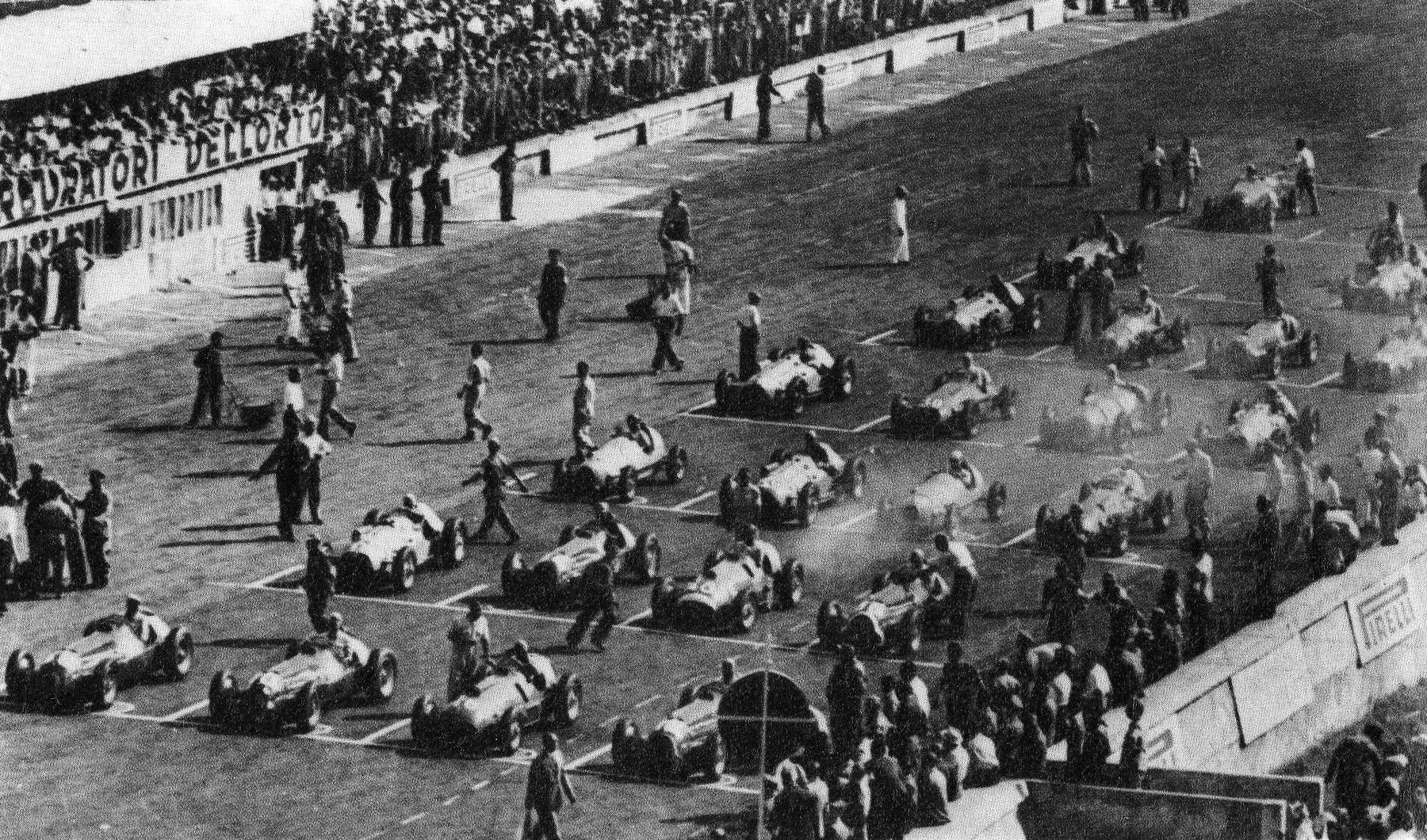 A high view over a grid of front-engined racing cars in staggered rows, drivers and mechanics standing among them; a packed grandstand at the top left behind a banner for Dell’Orto carburettors, and a Pirelli sign on the wall at the right.