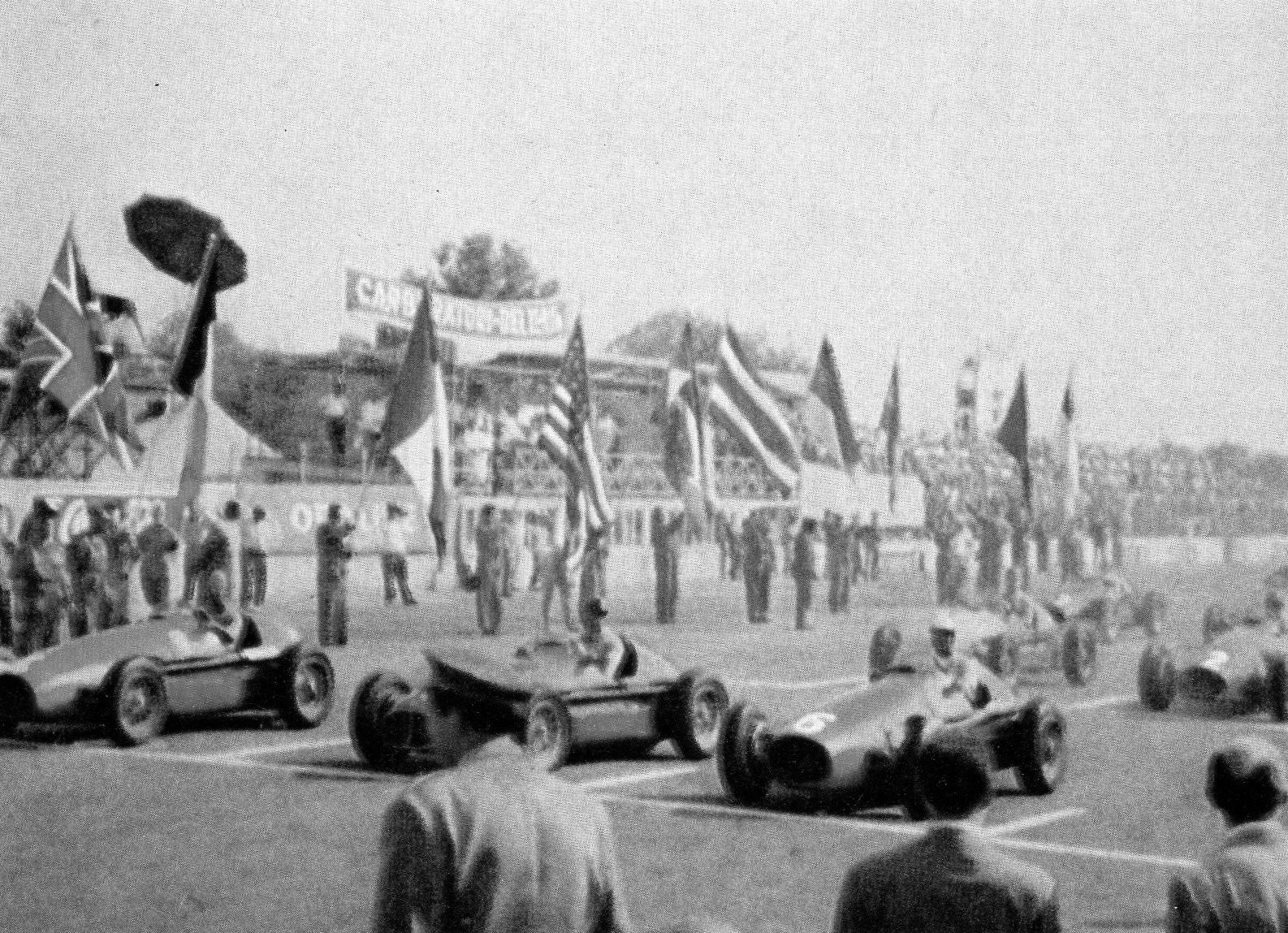 A faded print: front-engined racing cars on a grid under a row of national flags, drivers in the cockpits and mechanics among them, a grandstand behind and a man in a cap in the foreground.