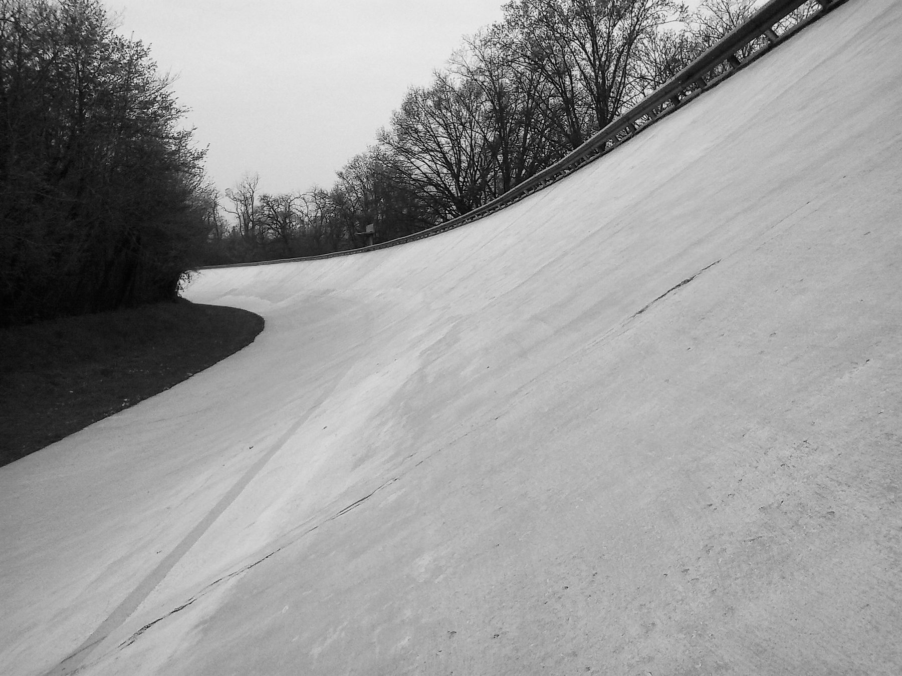 The steep concrete curve of a banked track sweeping up and away under an overcast sky, a painted line following the turn, a steel barrier along the rim and bare trees beyond.