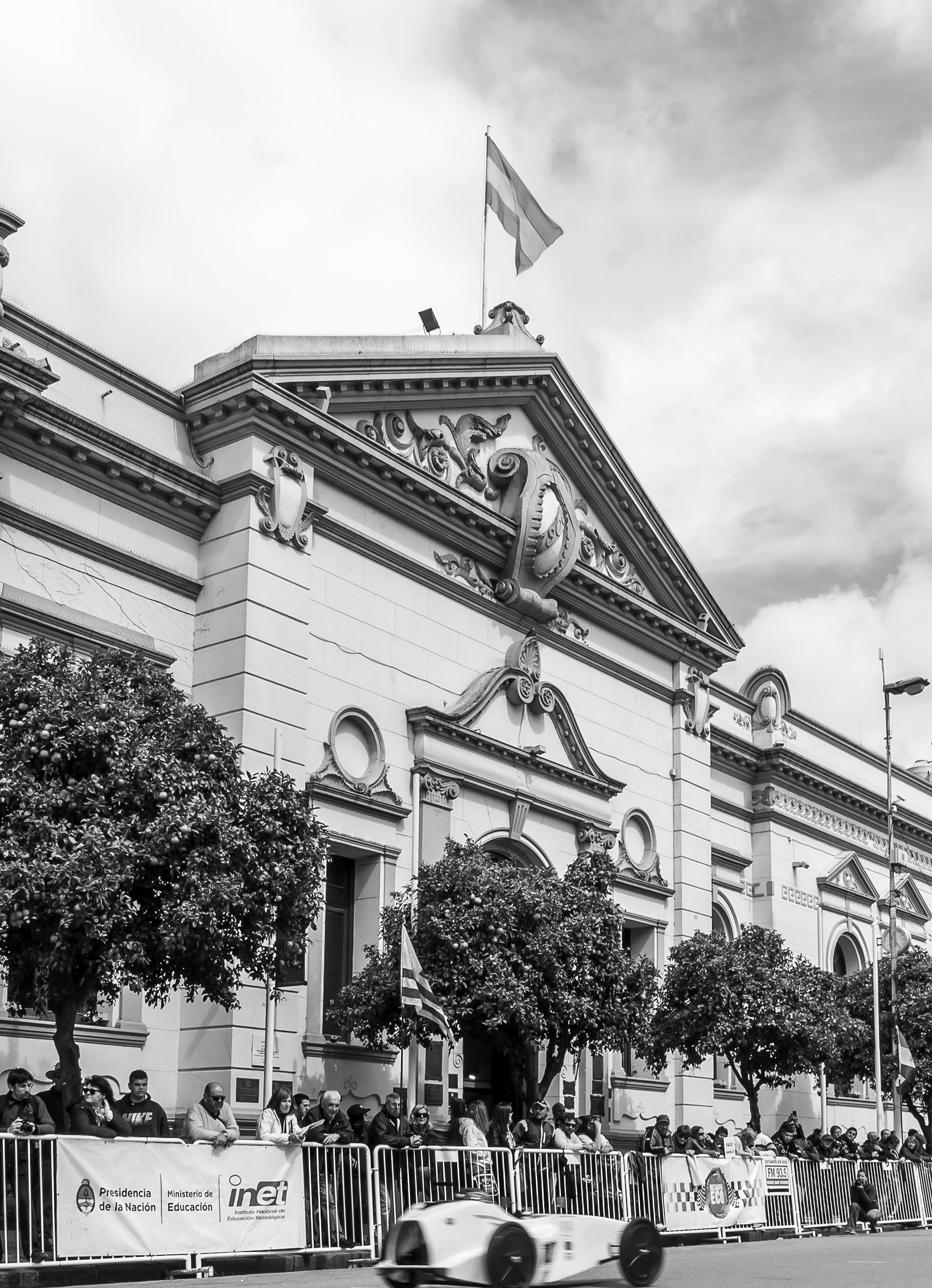The museum’s façade from the street on a bright day, a flag on the roof and flags at the door; spectators behind barriers hung with banners watch a small, low, streamlined electric racing car pass in the road.