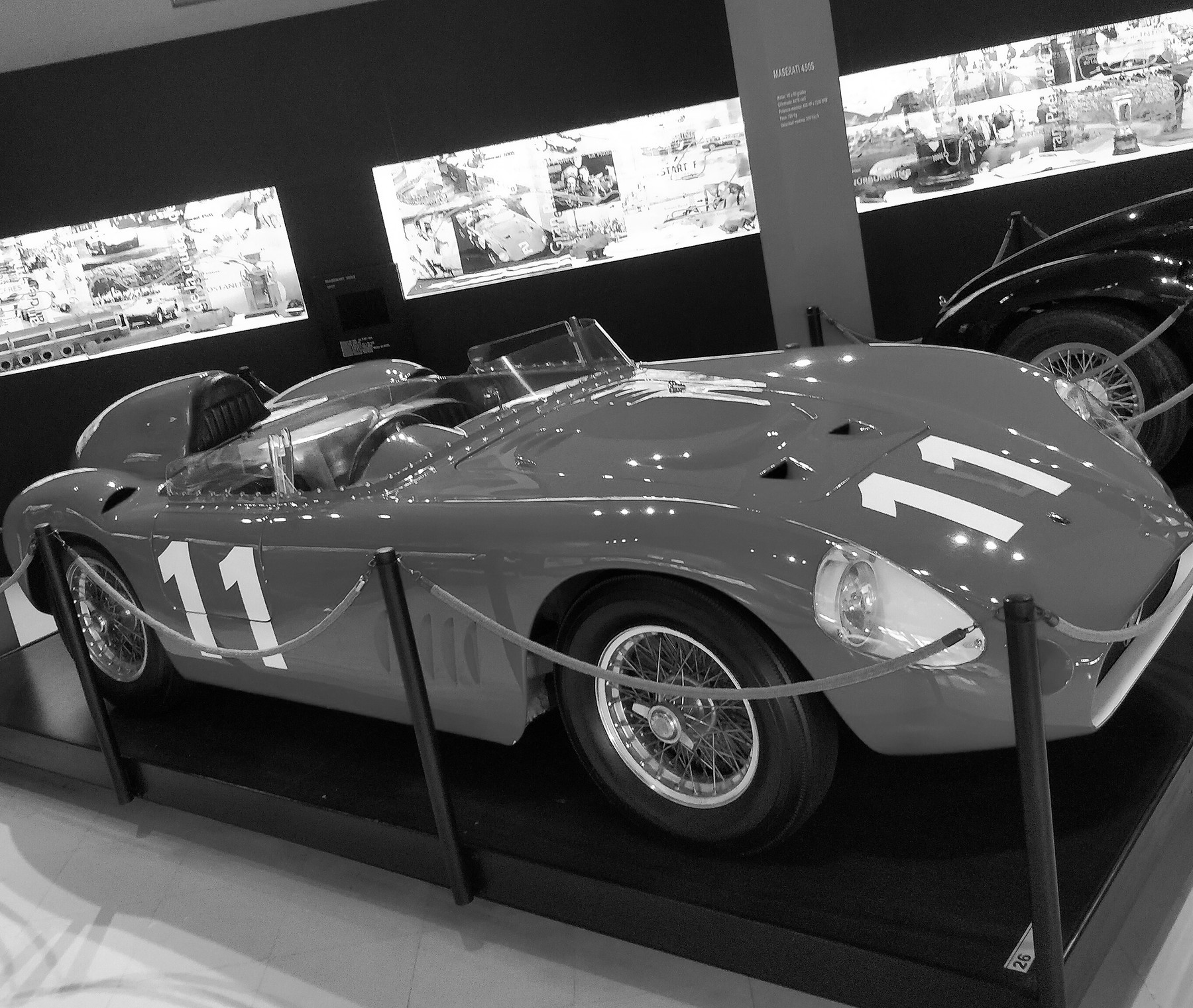 A low open sports-racing car numbered 11, with a small windscreen and wire wheels, roped off on a dark plinth in a museum; the tail of a dark car numbered 2 at the right and a lit cabinet of photographs behind.