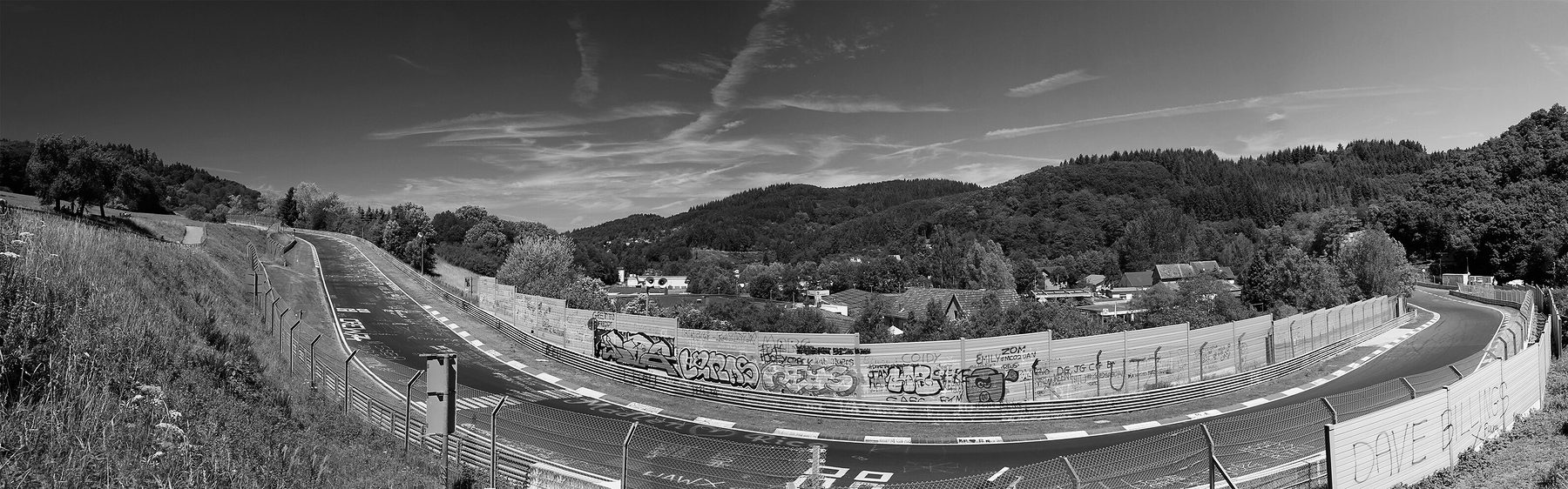 A wide panorama: a circuit sweeps down past walls covered with graffiti and steel barriers into a valley of wooded hills and houses, under a sky with vapour trails.