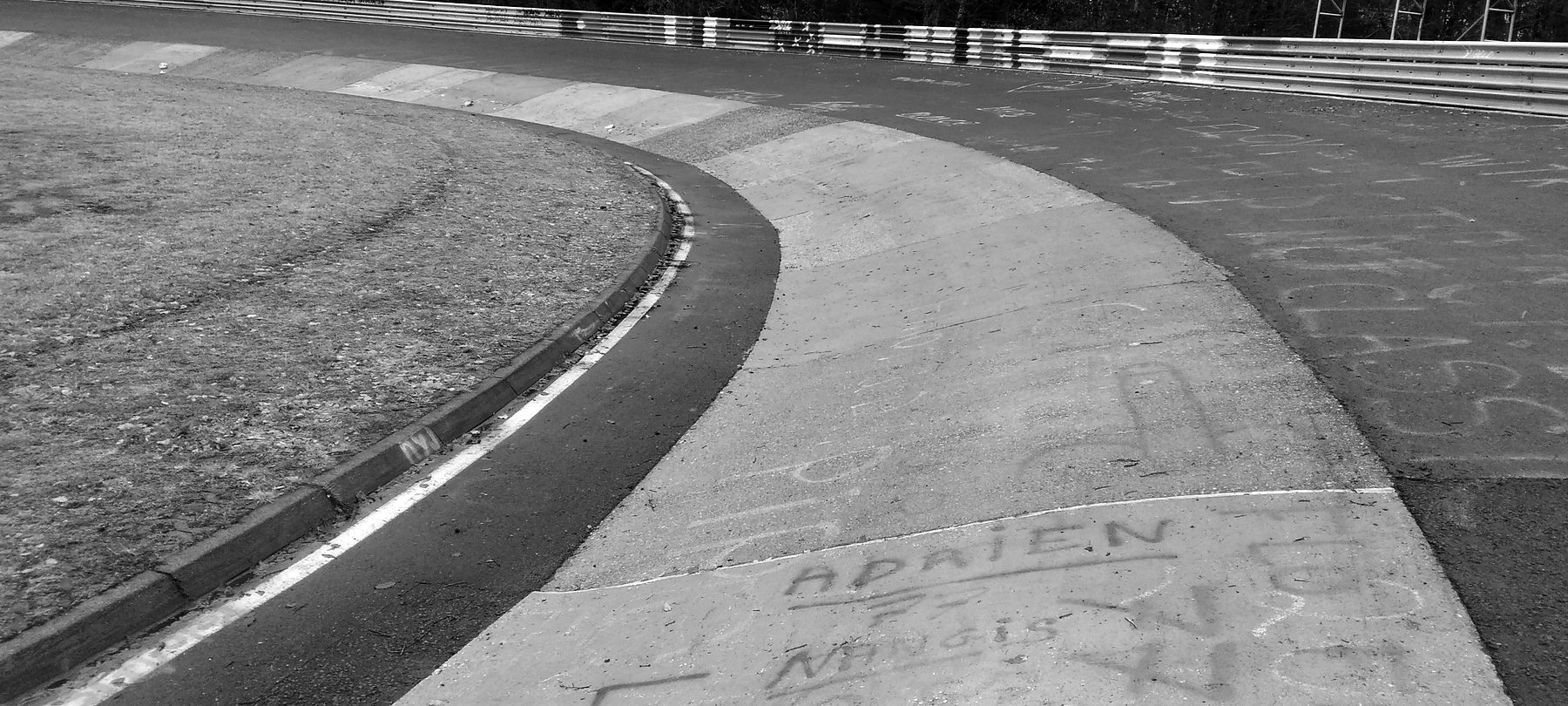The concrete banking of a tight hairpin curving away round a grassy infield, its slabs scrawled with names in chalk and paint, a crash barrier along the top edge.