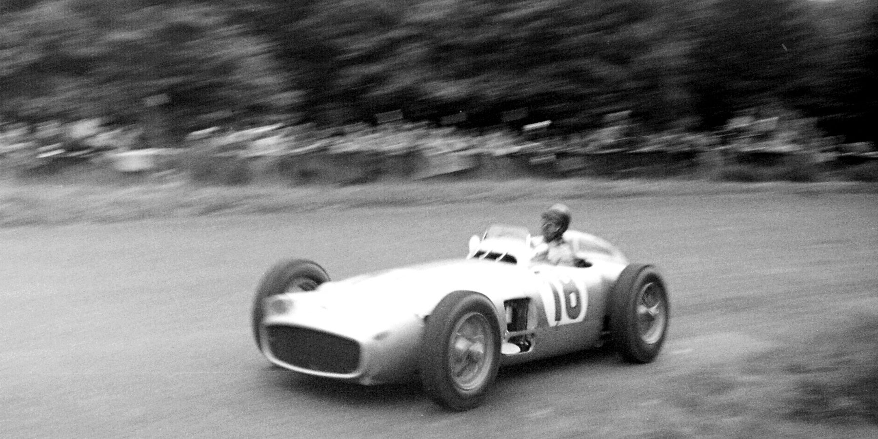 A panned photograph: Fangio, head and shoulders above the cockpit, sweeps round a bend in the open-wheeled Mercedes-Benz W196 numbered 18, trees and a line of spectators blurred into streaks behind.