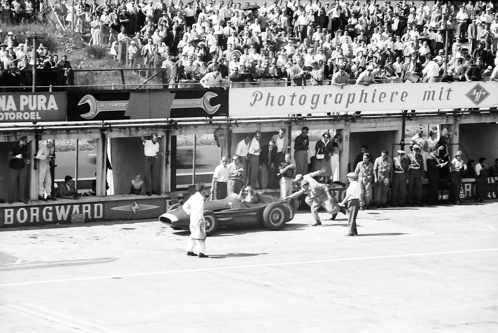 A racing car pulls away from a pit, a mechanic running beside it with an arm out; a man in white overalls stands in the foreground, and a packed stand and hoardings for Agfa and Borgward rise behind.
