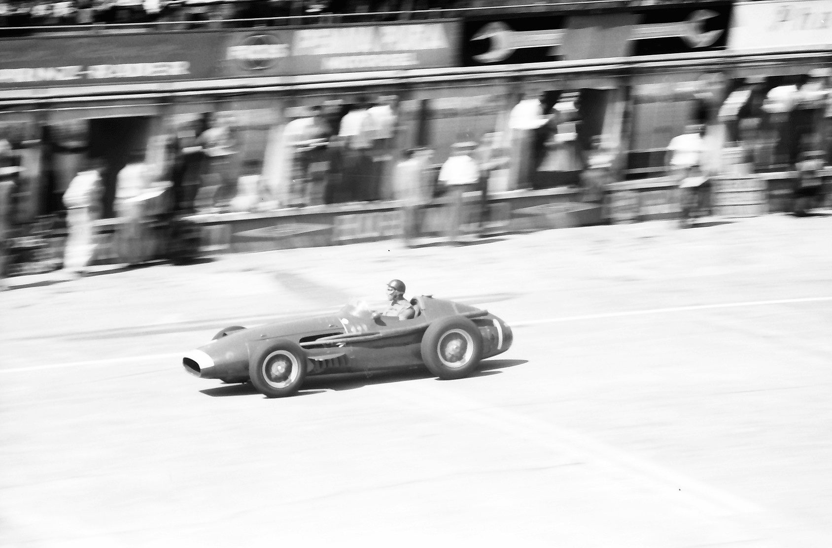 A panned photograph along the pits: Fangio crouches over the wheel of a Maserati 250F at speed, the pit wall and the people in front of it streaked into blurs behind.