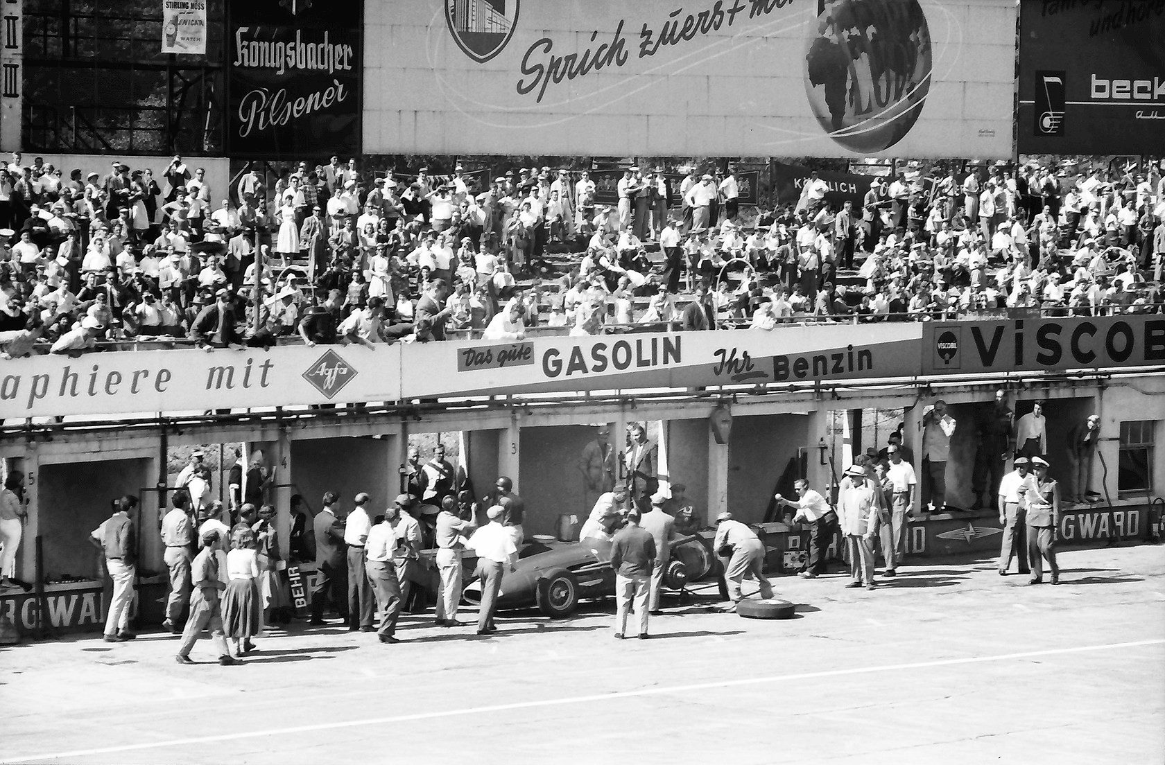 From the grandstand: mechanics crowd round a racing car at a pit under hoardings for Gasolin, Viscobil and Agfa, a wheel lying on the ground beside it; a packed stand above.