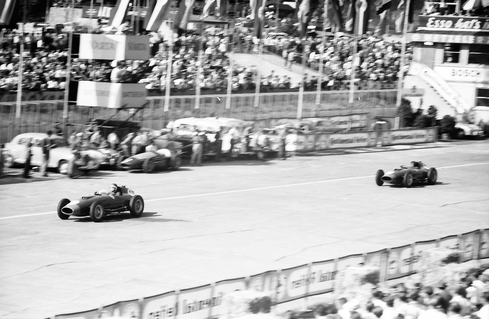 Two racing cars far apart on the start-and-finish straight, one close to the camera and one in the distance, past the pits, parked cars and a crowded stand with an Esso sign.