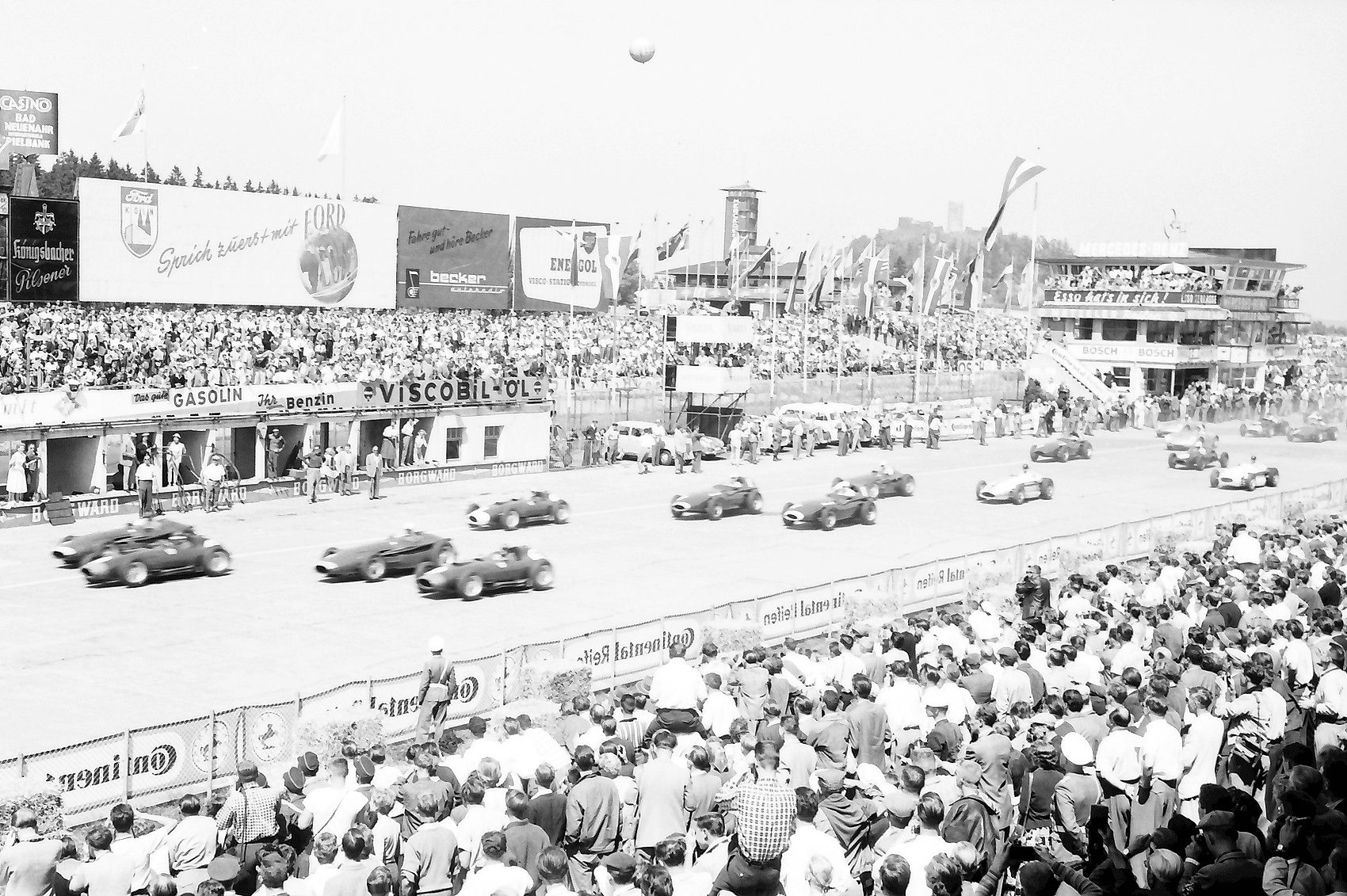 From the grandstand side: a field of front-engined racing cars pulls away along the start-and-finish straight, packed stands and a crowd in the foreground, advertising hoardings for Ford, Gasolin and Viscobil along the pits, the timing tower beyond.