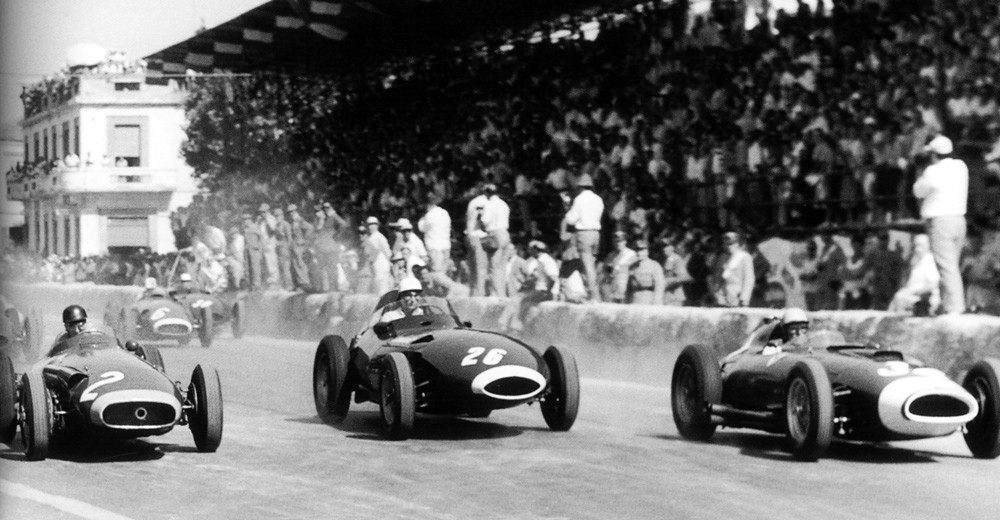 The front row leaves the line in front of a packed grandstand: a racing car numbered 2 on the left, another numbered 26 in the middle and a third on the right, smoke rising from their wheels.