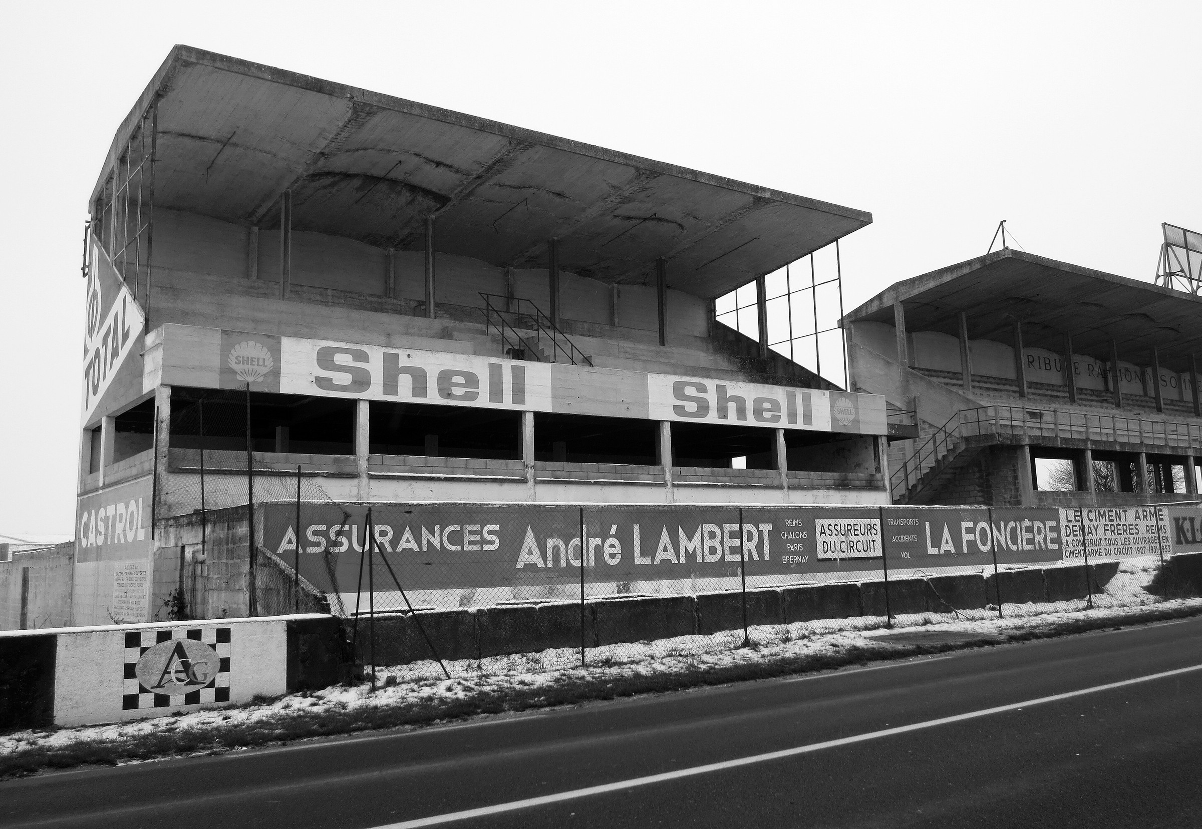 The empty concrete main grandstand of an old road circuit under its cantilevered roof, painted boards for Shell, Total and Castrol above a long banner for an insurer, snow on the verge and a dark road in front.