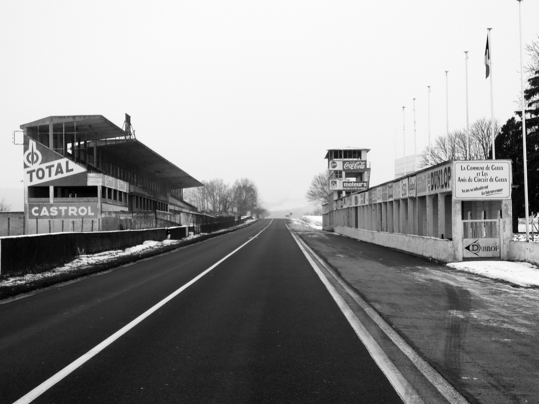 Looking down an empty straight between a grandstand on the left and a row of pits with painted advertisements on the right, a timing tower and flagpoles beyond, the road fading into winter haze.