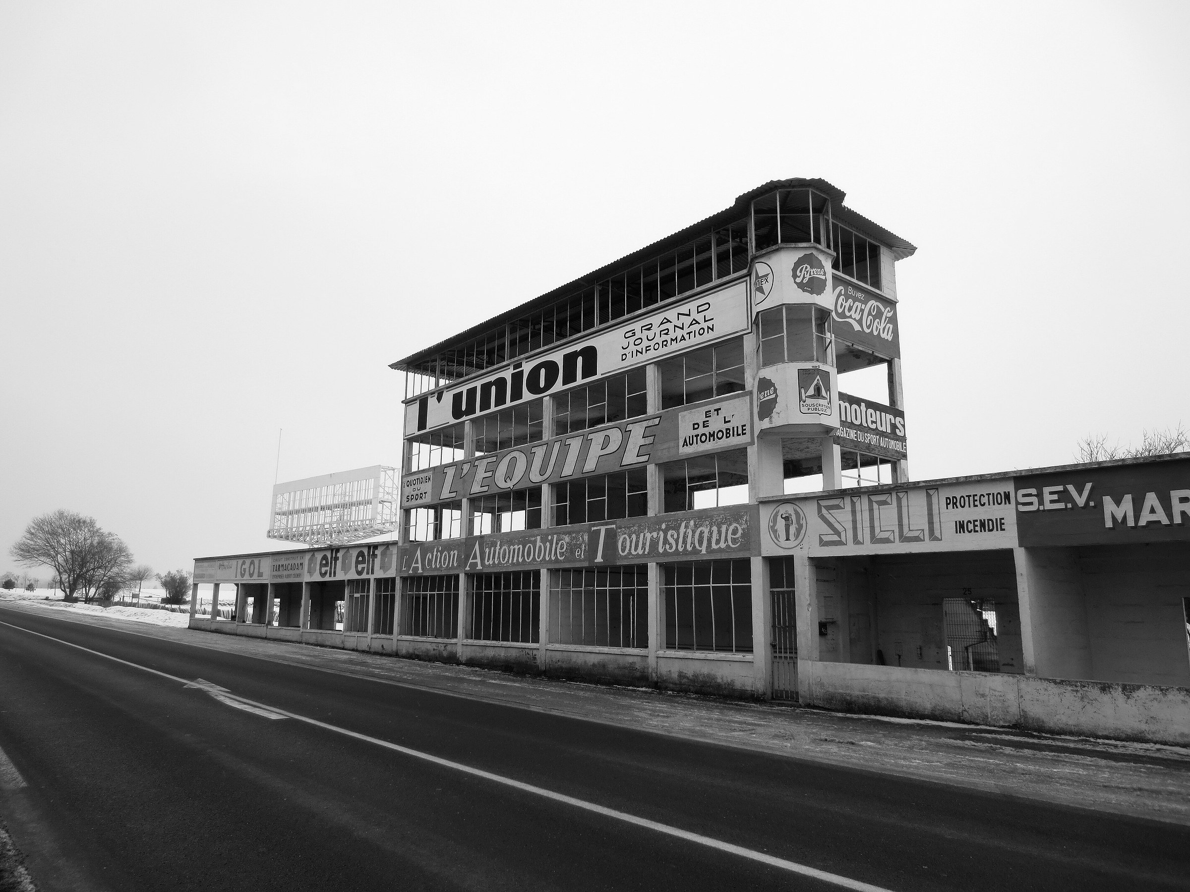 The preserved pits and multi-storey timing tower of an old road circuit beside an empty road on a grey, snowy day, painted with period advertisements for newspapers, motoring magazines and drinks.