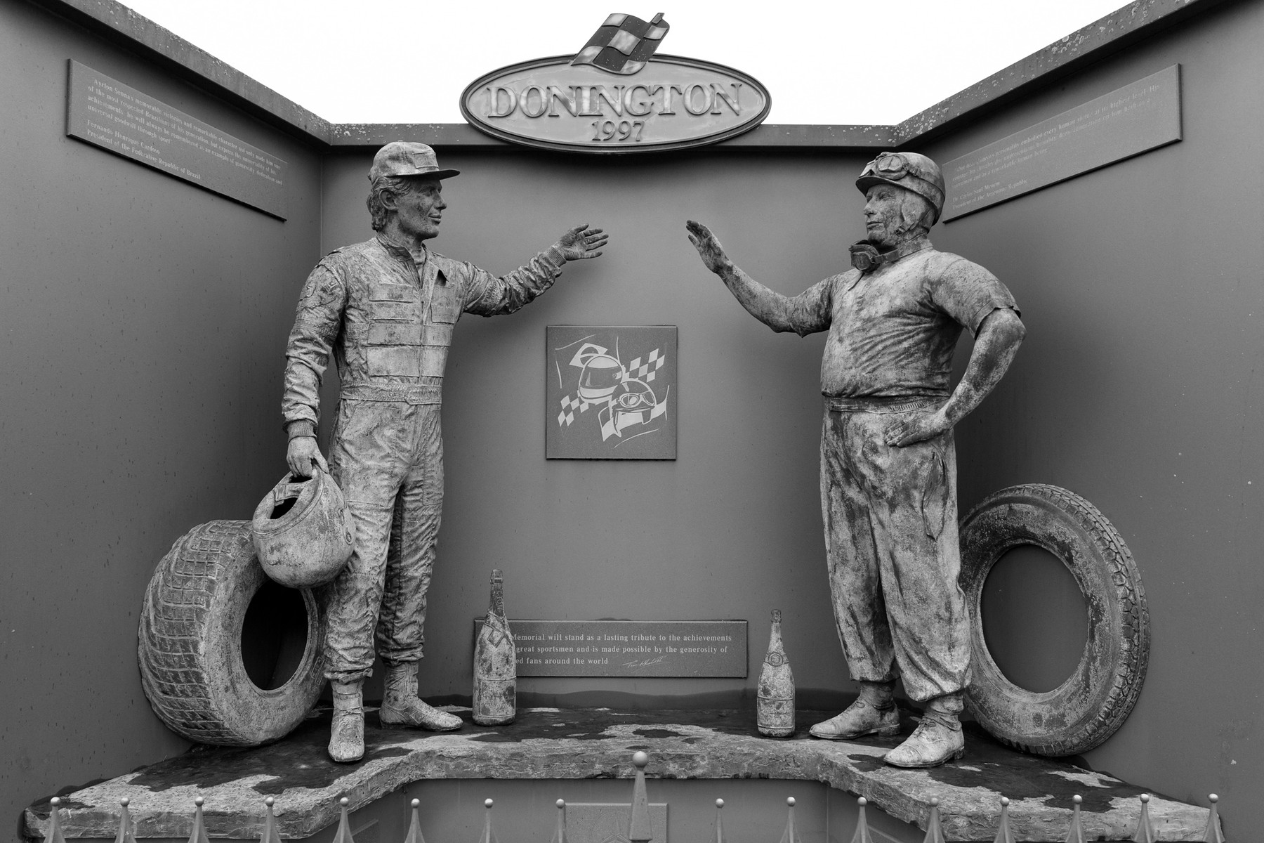 Two bronze figures in a three-sided grey niche under an oval sign reading Donington 1997: on the left a driver in overalls with a helmet under his arm and a tyre behind him, on the right a driver in shirt and baggy trousers with goggles round his neck and a hand on his hip; each raises a hand to the other, and bronze bottles and plaques stand between them.