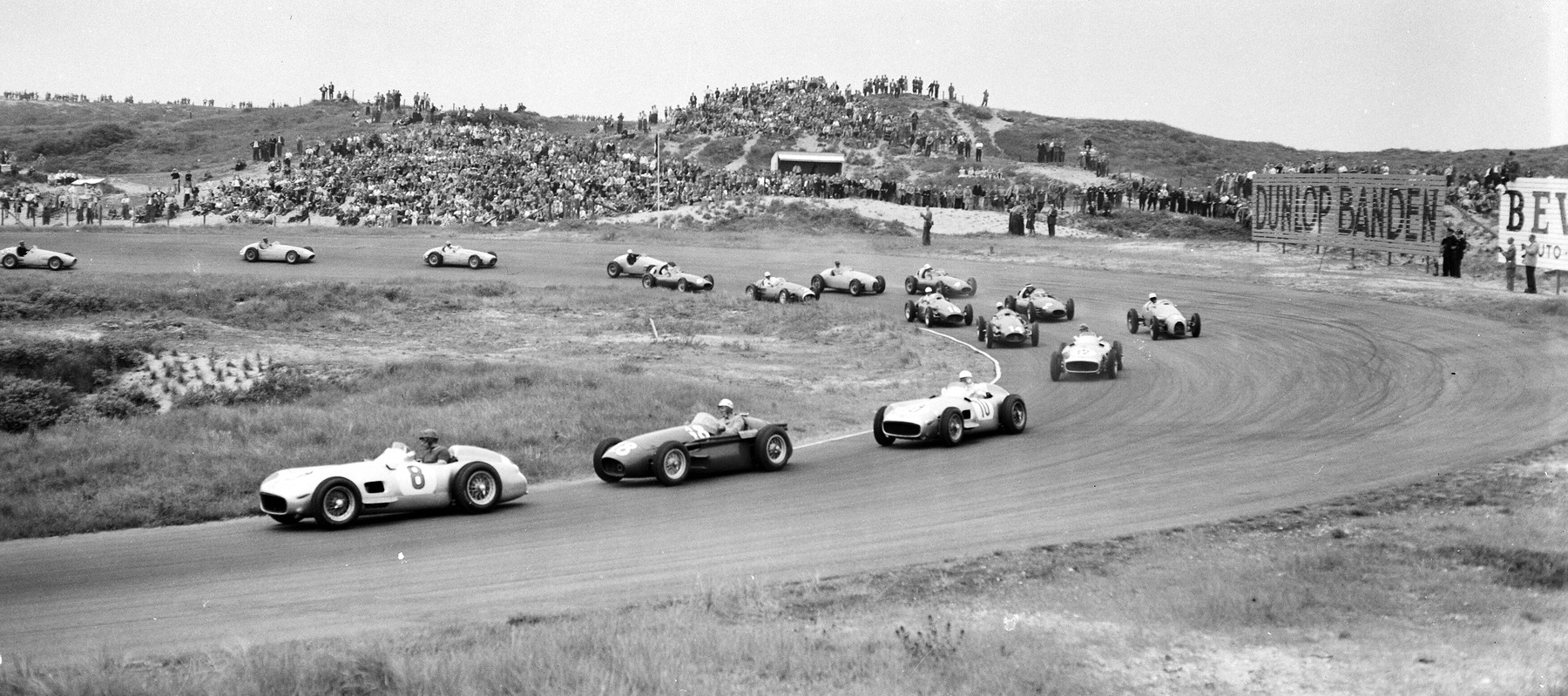 A wide view across the dunes: a string of front-engined racing cars rounds a long curve, a Mercedes-Benz numbered 8 in the lead, a Maserati numbered 18 and a second Mercedes-Benz numbered 10 behind it, spectators massed on the sandhills and a Dunlop hoarding at the right.